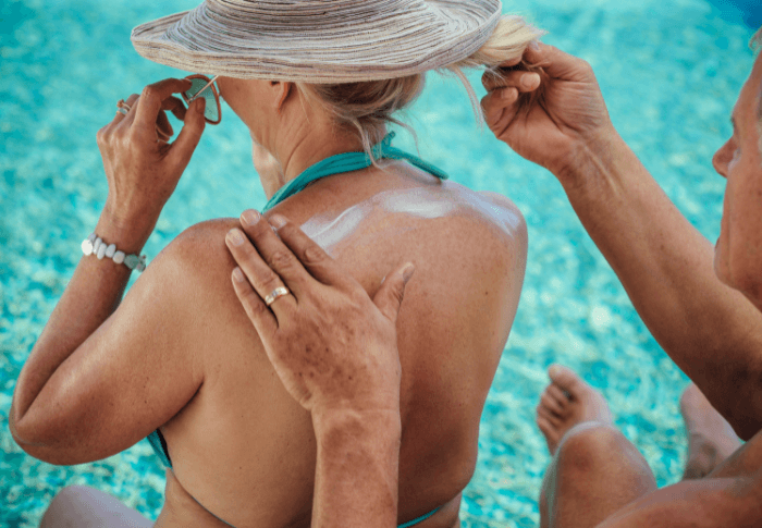 Your Guide to Staying Skin Safe This Summer Feature Image Woman wears hat at pool while man applies sunscreen to her back